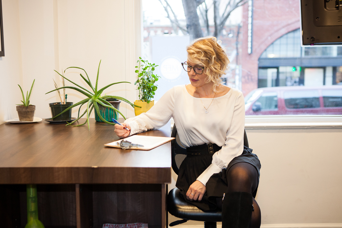 Image of MFP Founder and Managing Agent Sitting at a Table with a Pen Looking Down on a Clipboard with a Large Window Behind Her Showing the Partial Backdrop of a Brooklyn Street.