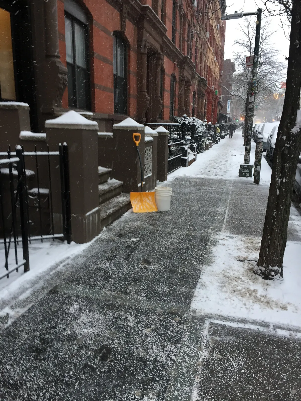 Snow-covered Brooklyn sidewalk outside brownstone buildings with a shovel and bucket and a section of the sidewalk shoveled for winter property upkeep.