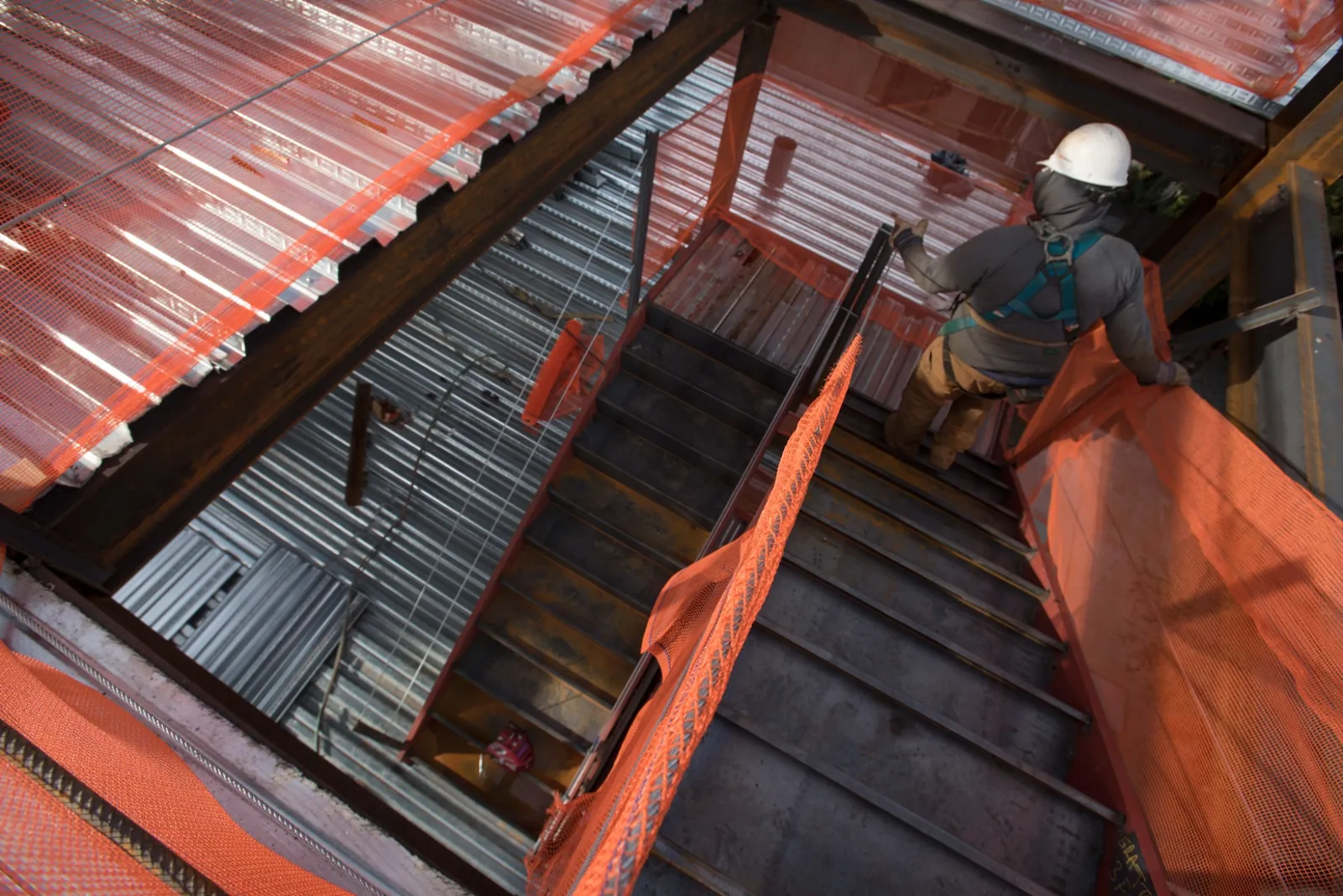 Image of a construction industry professional in a white safety helmet walking down raw concrete stairs with orange netting draped across open cavity spaces and steel beams and subsurface flooring material visible depicting a development project in a construction phase.