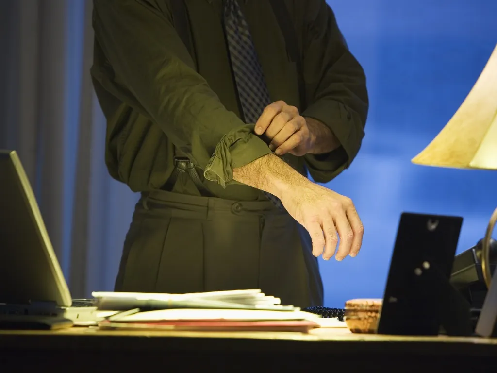 Landlord in a dress shirt and tie rolling up a sleeve beside a desk and laptop.