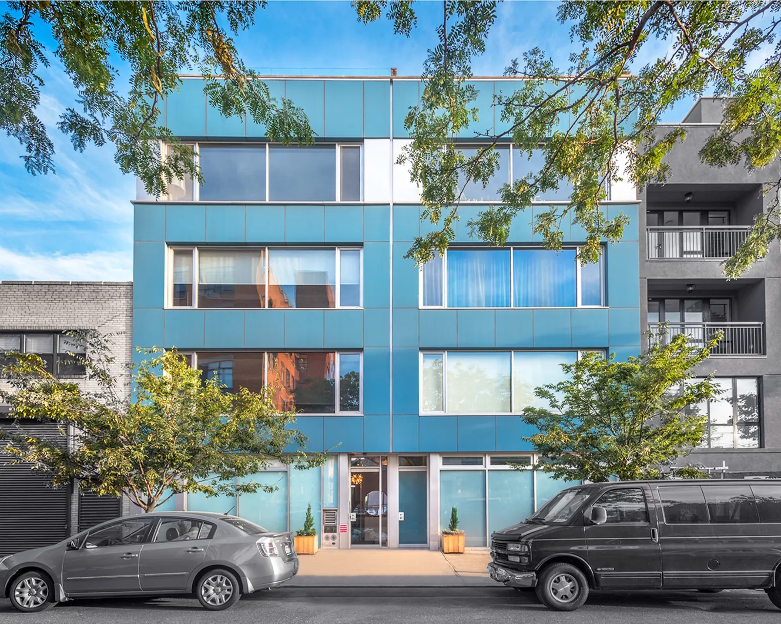 Brand new modern style building featuring reflective panels, large window spaces, and teal facade with trees and greenery visible in the image as well as parked vehicles on a NYC street.